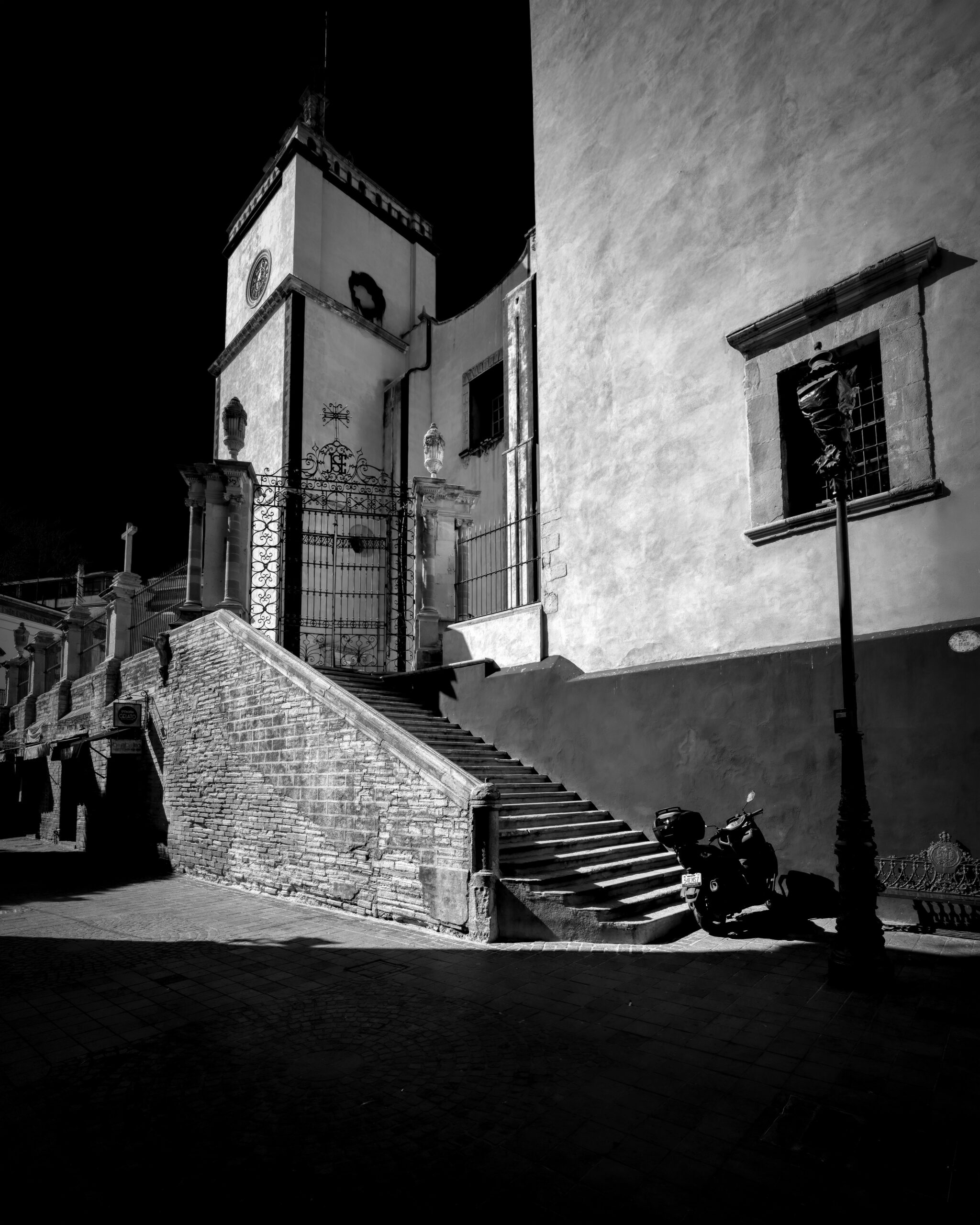 A sunlit Baroque church staircase and bell tower cut against deep shadow, a scooter parked below